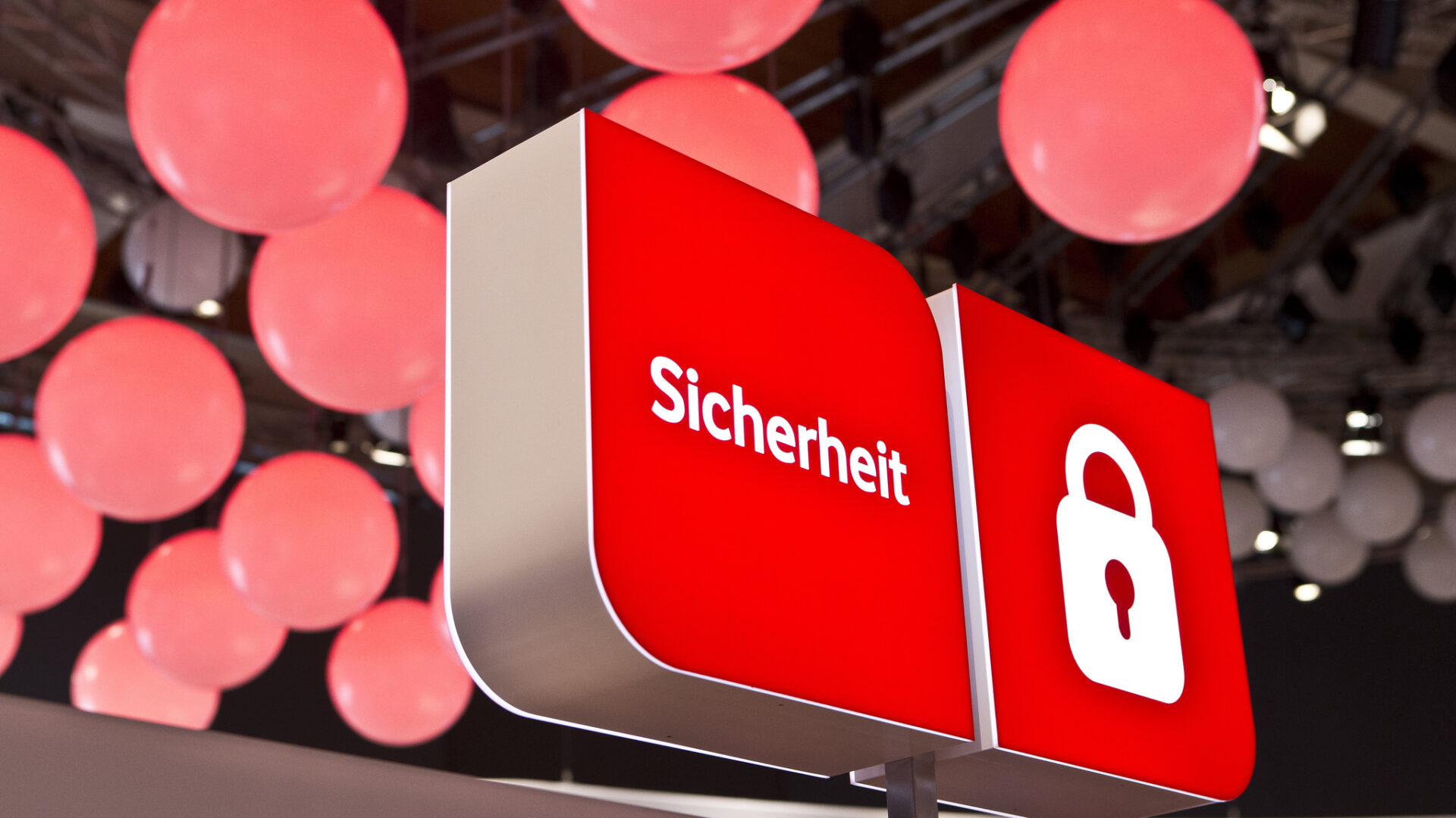Rotes Schild mit weißem Text „Sicherheit“ und einem weißen Vorhängeschloss-Symbol, im Hintergrund viele große, leuchtende, rosafarbene Kugeln an der Decke.