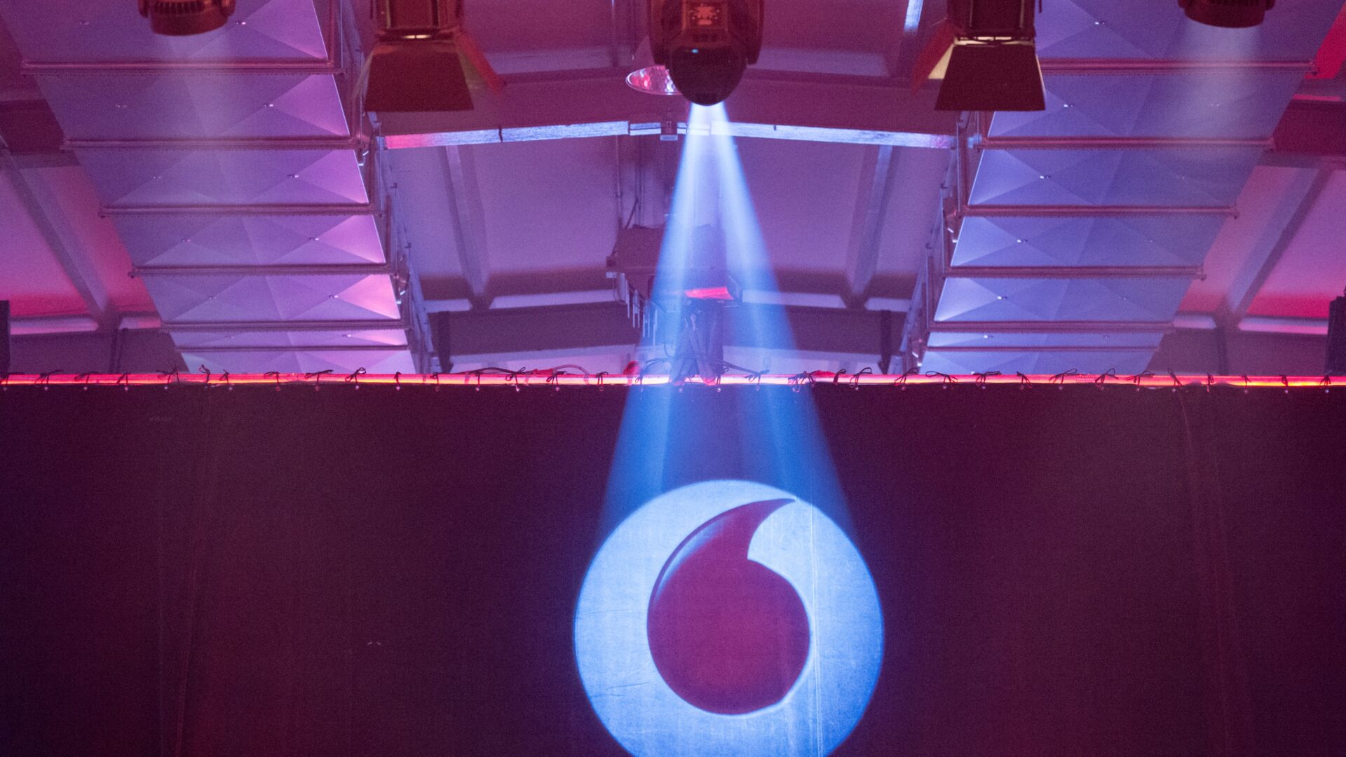 Beleuchtete Bühne mit Spotlights, auf der das Vodafone-Logo groß an eine Wand projiziert wird.