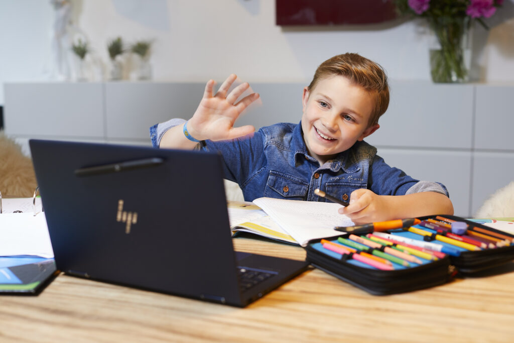 Ein Junge sitzt an einem Tisch mit einem Laptop, einem Notizbuch und einem Federmäppchen voller Stifte. Er trägt ein Jeanshemd, lächelt und winkt in die Richtung des aufgeklappten Laptops. Die Umgebung ist modern und freundlich eingerichtet.