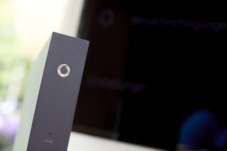 Nahaufnahme eines modernen grauen Geräts, vermutlich eines Modems oder Routers, mit dem Vodafone-Logo auf der Vorderseite. Im Hintergrund ist ein unscharfer Bildschirm oder Monitor zu sehen.