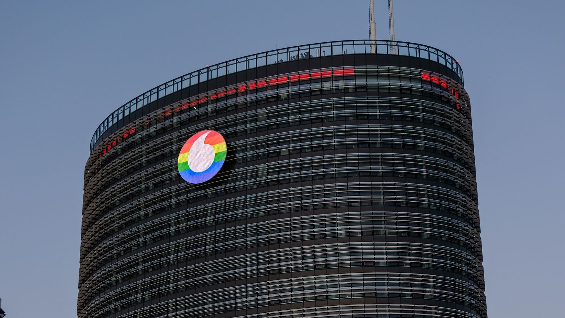 Der Tower des Vodafone Campus in Düsseldorf bei Dämmerung, auf dessen Fassade das Vodafone-Logo in Regenbogenfarben projiziert ist. Das Gebäude hat eine runde Form und ist von einem Abendhimmel umgeben. Vor dem Gebäude sind einige Bäume zu sehen.