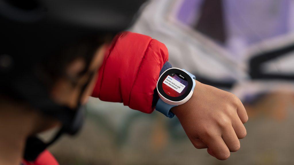 Ein Kind mit roter Jacke trägt eine Smartwatch am Handgelenk, die eine Nachricht anzeigt. Der Hintergrund ist unscharf, und das Kind scheint einen Helm zu tragen, was auf eine Aktivität im Freien hindeutet.