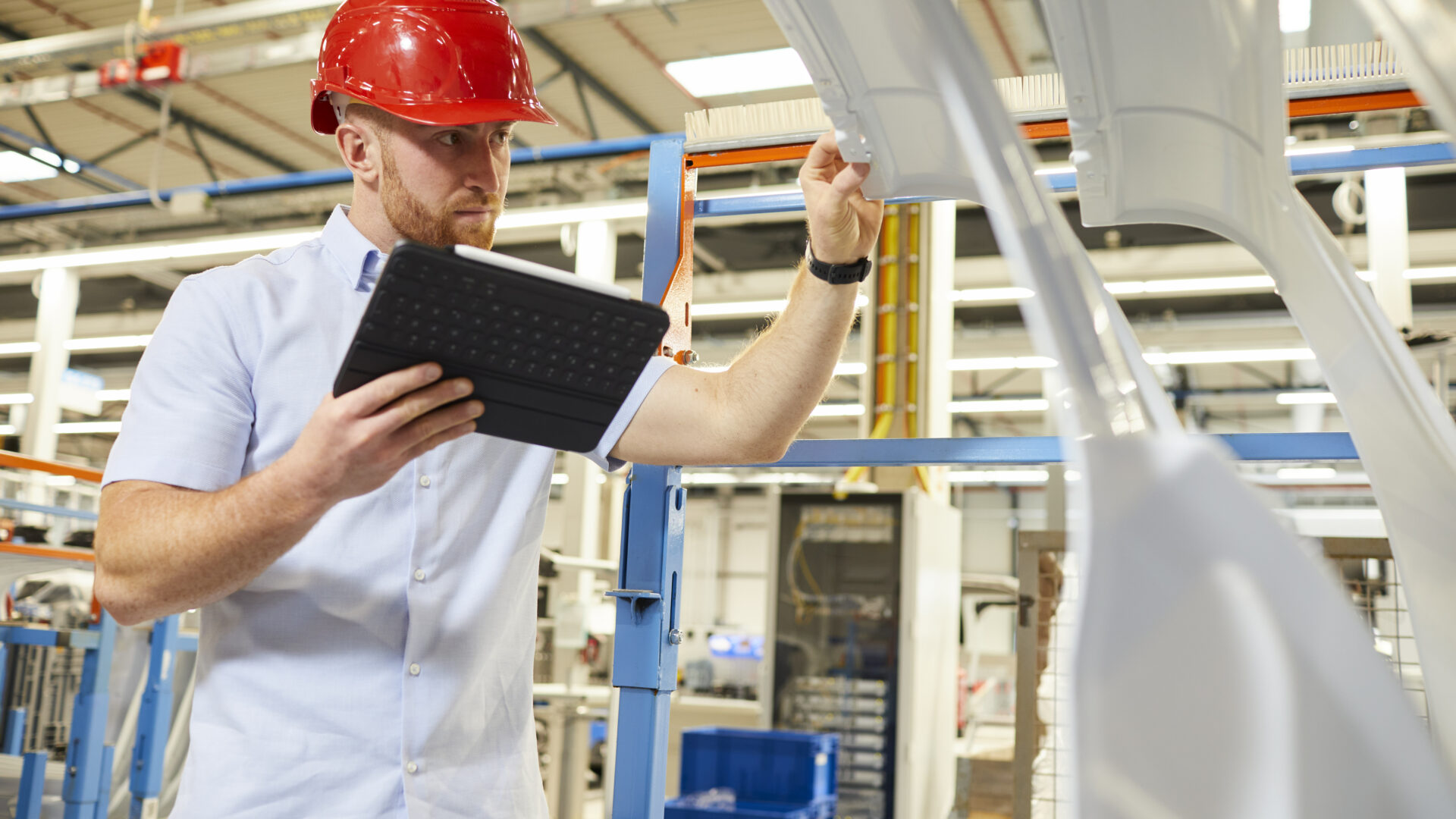 Das Bild zeigt einen Mann in einem industriellen oder Fertigungsumfeld. Er trägt einen roten Sicherheitshelm und ein helles Hemd. Der Mann betrachtet ein großes, weißes Objekt, möglicherweise ein Teil einer Maschine oder eines Fahrzeugs. In der anderen Hand hält er ein Tablet, vermutlich zur Datenerfassung oder zur Überprüfung von Spezifikationen. Der Hintergrund zeigt eine Fabrikhalle mit blauen und weißen Strukturen, Regalen und weiteren industriellen Ausstattungen.