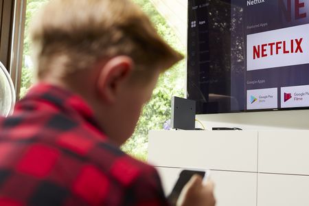 Ein Raum mit einem großen Flachbildfernseher, der an der Wand montiert ist. Der Bildschirm zeigt Streaming-Dienste wie Netflix und Prime Video sowie Symbole für Apps wie YouTube. Ein Kind in einem rot-schwarz karierten Hemd hält eine Fernbedienung. Der Raum ist hell, mit Licht von einem Fenster. Unter dem Fernseher steht ein weißes Möbelstück.