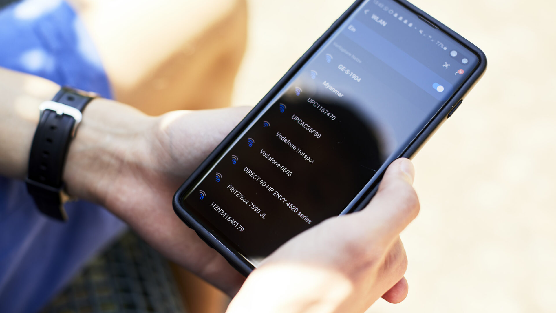 Das Bild zeigt eine Person, die ein Smartphone mit beiden Händen hält. Auf dem Bildschirm des Telefons ist eine Anwendung mit einer Liste von Einstellungen oder Optionen zu sehen. Die Benutzeroberfläche ist im Dunkelmodus, mit blauem und weißem Text auf einem dunklen Hintergrund. Die Person trägt eine schwarze Smartwatch am Handgelenk. Der Hintergrund ist leicht verschwommen, was darauf hindeutet, dass die Person möglicherweise draußen oder in einem gut beleuchteten Bereich sitzt. Der Fokus liegt auf dem Smartphone-Bildschirm, was auf eine Interaktion mit dem Gerät hinweist.