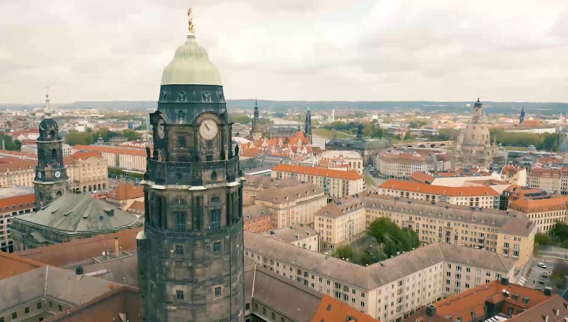 Luftaufnahme der Stadt Dresden mit Blick auf den Turm des Dresdner Rathauses im Vordergrund. Im Hintergrund sind die Frauenkirche und weitere historische Gebäude zu sehen. Die Szene zeigt eine Mischung aus alten und modernen Architekturmerkmalen in einer städtischen Landschaft.