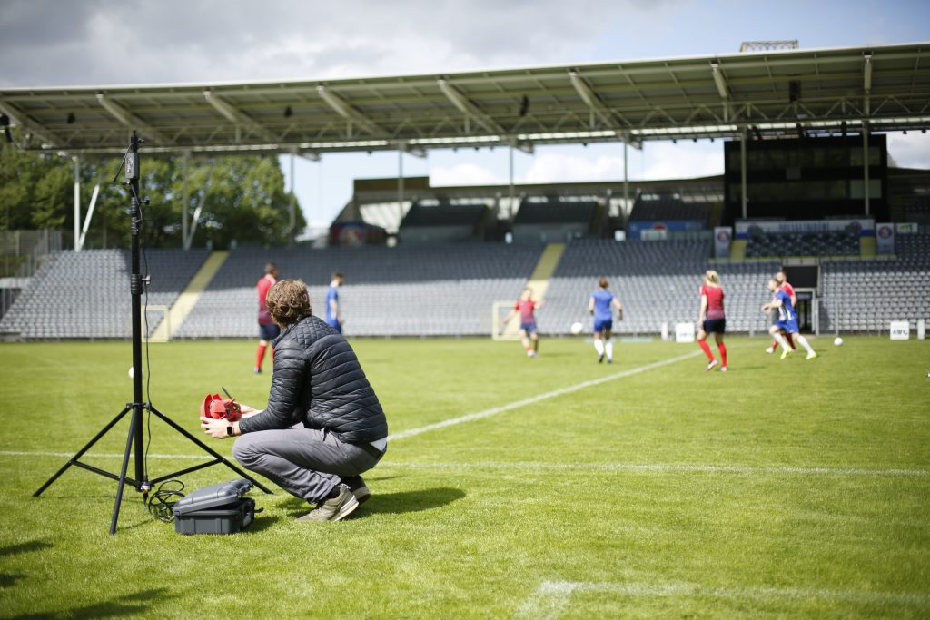 Eine Person sitzt auf einem Fußballfeld neben einem Stativ und beobachtet ein Spiel. Im Hintergrund sind mehrere Fußballspieler in roten und blauen Trikots sowie leere Tribünen zu sehen. Das Wetter ist bewölkt, aber hell.