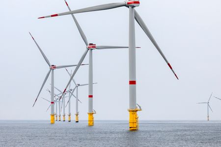Mehrere Offshore-Windkraftanlagen stehen im Meer, mit weißen Rotorblättern und roten Markierungen. Die Fundamente der Windräder sind gelb und ragen aus dem Wasser. Der Himmel ist bewölkt und das Meer wirkt ruhig.