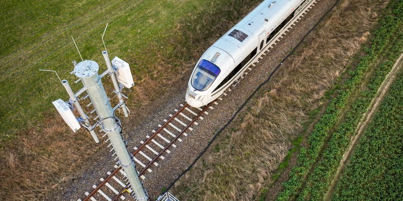 Luftaufnahme eines weißen Hochgeschwindigkeitszuges, der auf einem einzelnen Gleis durch eine grüne, ländliche Landschaft fährt. Im Vordergrund steht ein Mobilfunkmast mit mehreren Antennen, der sich direkt neben den Bahngleisen befindet. Links und rechts der Schienen sind Gras- und Ackerflächen zu sehen. Das Bild vermittelt die Nähe moderner Zugtechnik und Telekommunikation in einer natürlichen Umgebung.