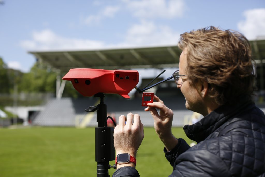 Eine Person installiert eine rote SIM-Karte in ein rotes Gerät mit Antennen, das auf einem Stativ montiert ist. Im Hintergrund sind unscharf Fußballspieler und leere Tribünen zu sehen.