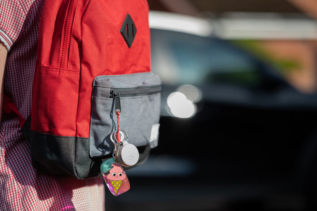 Ein roter Rucksack mit einer grauen Vordertasche, an der ein GPS-Tracker und ein Schlüsselanhänger mit einem niedlichen Design befestigt sind. Der Rucksack wird von einer Person getragen, die ein kariertes Hemd trägt. Im Hintergrund ist ein unscharfes Auto zu sehen.
