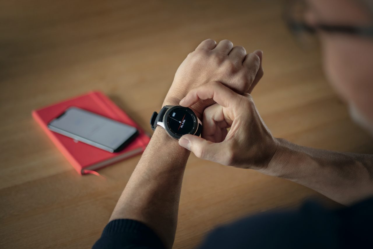 Eine Person überprüft eine Smartwatch am Handgelenk. Im Hintergrund liegen ein Smartphone und ein rotes Notizbuch auf einem Holztisch.