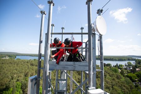 Zwei Personen in roter Schutzkleidung und schwarzen Helmen arbeiten an einem Mobilfunkmast. Sie sind mit Sicherheitsausrüstung gesichert und führen Arbeiten in großer Höhe aus. Im oberen Bereich des Mastes ist eine Satellitenschüssel montiert. Im Hintergrund erstreckt sich eine Landschaft mit Wäldern und einem großen See. Der Himmel ist größtenteils klar mit wenigen Wolken.