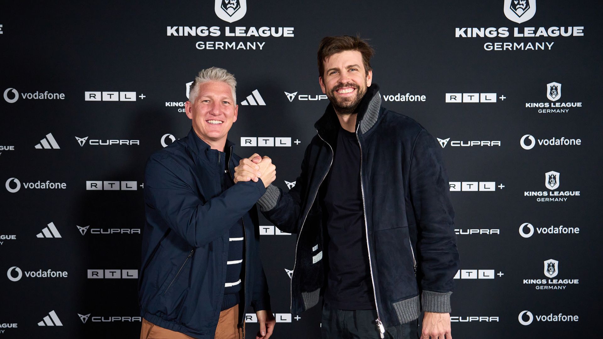 Bastian Schweinsteiger und Gerard Piqué stehen vor einer Sponsorenwand mit Logos der "Kings League Germany" und verschiedenen Partnern wie Vodafone, RTL, und Adidas. Sie geben sich die Hand und posieren für ein Foto. Die Szene vermittelt den Eindruck einer sportlichen oder geschäftlichen Veranstaltung, bei der Partnerschaften und Kooperationen im Vordergrund stehen.