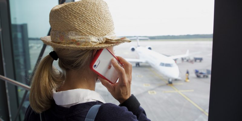 Eine Person mit Strohhut und Rucksack telefoniert mit einem Smartphone und schaut durch ein Fenster auf ein Flugzeug, das auf dem Rollfeld eines Flughafens steht.

