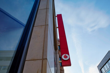 Außenansicht eines Gebäudes mit rotem Vodafone-Schild an der Fassade und blauem Himmel im Hintergrund.