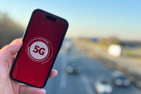Eine Hand hält ein Smartphone, dessen Bildschirm ein rotes 5G-Symbol zeigt. Im Hintergrund ist eine Autobahn mit mehreren fahrenden Autos zu sehen, die durch eine ländliche Gegend führt. Der Himmel ist klar und blau, was eine helle und offene Atmosphäre schafft. Die Szene symbolisiert die Verbindung von moderner Technologie mit Mobilität und Infrastruktur.