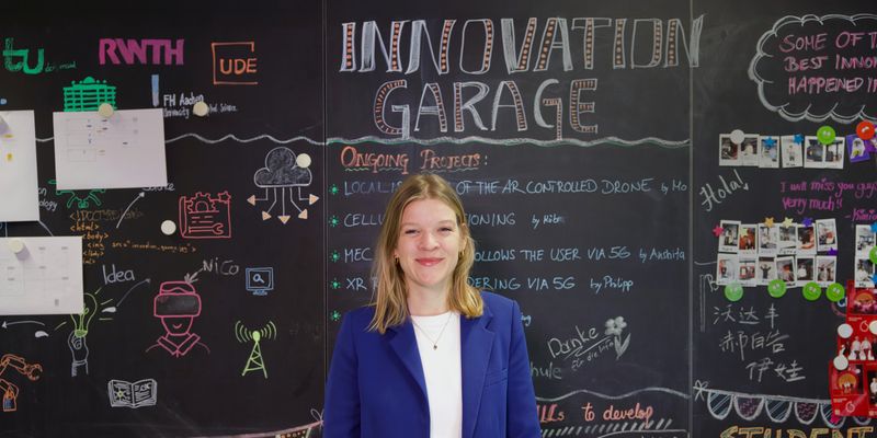 Laura Biermann steht vor einer kreativ gestalteten Wand, die mit dem Titel "Innovation Garage" beschriftet ist. Die Wand ist mit verschiedenen Zeichnungen, Notizen und Ideen versehen, die auf ein kreatives und inspirierendes Arbeitsumfeld hinweisen. Sie trägt einen blauen Blazer.