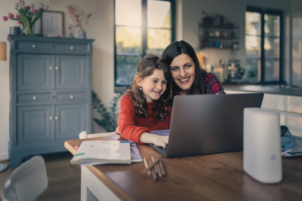 Zwei Personen sitzen an einem Tisch und arbeiten gemeinsam an einem Laptop. Auf dem Tisch liegen Bücher und Stifte. Der Raum ist hell und gemütlich eingerichtet, mit einem großen Fenster und einem Schrank im Hintergrund.