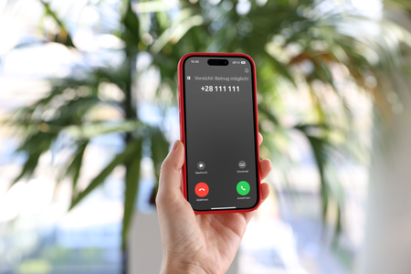 Eine Person hält ein rotes Smartphone in der Hand und betrachtet den Bildschirm. Auf dem Display ist ein eingehender Anruf von einer unbekannten Nummer zu sehen, begleitet von dem Hinweis "Vorsicht Betrug möglich". Die Umgebung ist hell und im Hintergrund sind unscharf grüne Pflanzen zu erkennen.