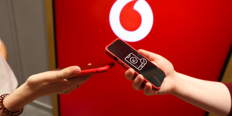 Zwei Personen halten jeweils ein rotes Smartphone und führen eine Aktion aus, die eine Datenübertragung oder eine mobile Zahlung darstellt. Im Hintergrund ist ein großes Vodafone-Logo auf einem roten Bildschirm sichtbar. Die Szene vermittelt den Eindruck von moderner Technologie und Konnektivität.
