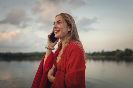 Eine Person steht am Ufer eines ruhigen Sees, in eine Jacke gehüllt und telefoniert. Im Hintergrund sind bewölkter Himmel und Bäume am anderen Ufer des Sees zu erkennen, die eine friedliche und entspannte Atmosphäre schaffen. Das Wasser des Sees ist ruhig und spiegelt die Umgebung wider.
