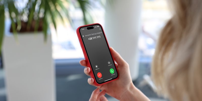 Eine Person hält ein rotes Smartphone in der Hand und betrachtet den Bildschirm. Auf dem Display ist ein eingehender Anruf von einer unbekannten Nummer zu sehen, begleitet von dem Hinweis "Vorsicht Betrug möglich". Die Umgebung ist hell und im Hintergrund sind unscharf grüne Pflanzen zu erkennen.