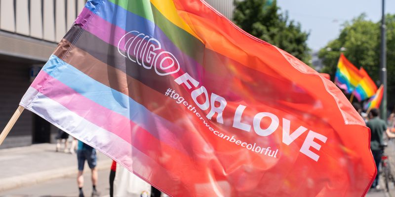 Eine Pride-Flagge mit erweiterten Farben für mehr Inklusion (Braun, Schwarz, Hellblau, Rosa und Weiß zusätzlich zu den Regenbogenfarben) weht im Wind auf einer Straße während einer Parade oder Demonstration. Auf der Flagge steht „GO FOR LOVE“ und darunter der Hashtag „#togetherwecanbecolorful“. Im Hintergrund sind weitere Regenbogenflaggen zu sehen, einige Menschen laufen auf der Straße, und rechts und links stehen Gebäude und Bäume.