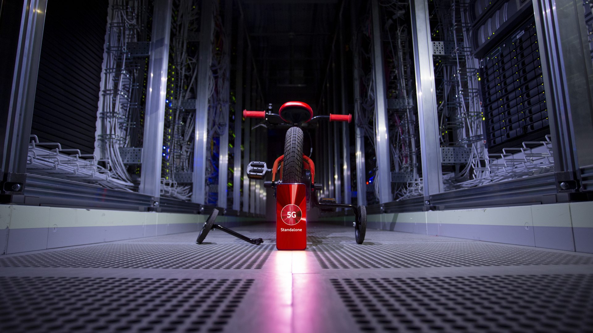 Ein rotes Kinderfahrrad steht in einem futuristisch anmutenden Raum, der von Servern und technischer Infrastruktur umgeben ist. Vor dem Fahrrad ist ein rotes Schild mit der Aufschrift "5G Vodafone" zu sehen. Die Beleuchtung ist gedämpft.