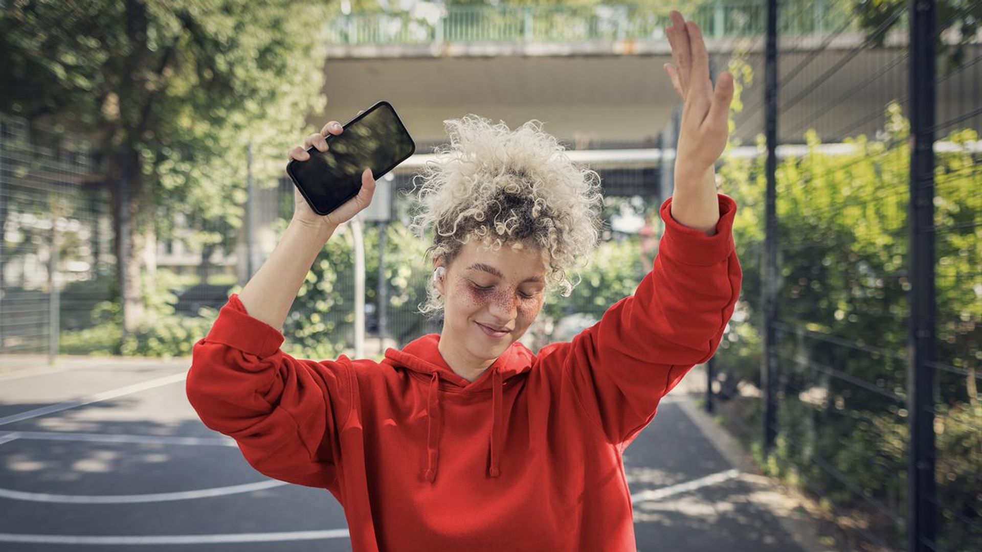 Eine Person in einem roten Hoodie steht auf einem Basketballplatz im Freien und hält ein Smartphone in der Hand. Sie scheint in Bewegung zu sein, möglicherweise beim Tanzen oder Jubeln. Die Umgebung ist von Bäumen und einem Zaun umgeben, was eine sportliche und lebendige Atmosphäre vermittelt.