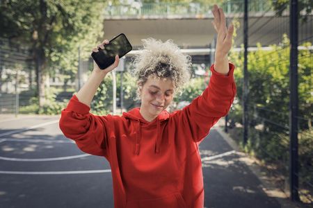 Eine Person in einem roten Hoodie steht auf einem Basketballplatz im Freien und hält ein Smartphone in der Hand. Sie scheint in Bewegung zu sein, möglicherweise beim Tanzen oder Jubeln. Die Umgebung ist von Bäumen und einem Zaun umgeben, was eine sportliche und lebendige Atmosphäre vermittelt.