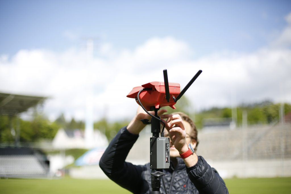 Eine Person justiert ein rotes Gerät mit Antennen auf einem Stativ auf einem Fußballfeld. Im Hintergrund sind leere Tribünen und ein bewölkter Himmel zu sehen.