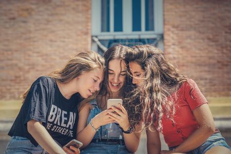 Drei junge Personen sitzen zusammen und schauen auf ein Smartphone, das eine von ihnen hält. Sie sind lässig gekleidet und scheinen sich in einer entspannten Umgebung im Freien zu befinden, möglicherweise vor einem Gebäude. Die Szene vermittelt den Eindruck von Freundschaft und gemeinsamer Zeit, während sie sich mit digitaler Technologie beschäftigen.
