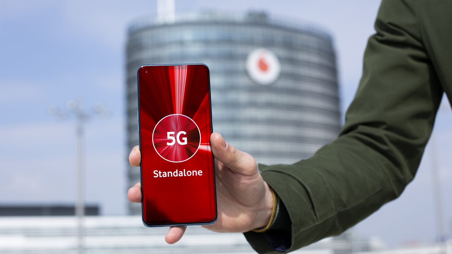 Eine Hand hält ein Smartphone, dessen Bildschirm die Worte "5G Standalone" auf einem roten Hintergrund zeigt. Im Hintergrund ist ein modernes Gebäude mit dem Vodafone-Logo zu sehen. Der Himmel ist blau und klar.