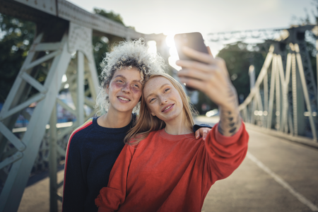 Zwei Personen stehen auf einer Brücke und machen ein Selfie mit einem Smartphone. Sie sind lässig gekleidet und genießen offensichtlich den Moment in der Abendsonne. Die Szene vermittelt den Eindruck von Freundschaft und Freude in einer urbanen Umgebung.