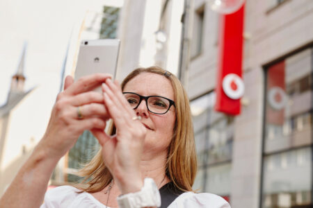Eine Frau mit langen, blonden Haaren und Brille hält ein Smartphone hoch und macht vermutlich ein Foto oder ein Selfie. Sie steht im Freien vor einem modernen Gebäude, an dem ein großes rotes Banner mit einem weißen Ring und Kreis hängt. Die Frau trägt ein weißes Oberteil und eine weiße Armbanduhr. Im Hintergrund sind Glasfassaden, spiegelnde Fenster und ein unscharfer Kirchturm zu sehen.