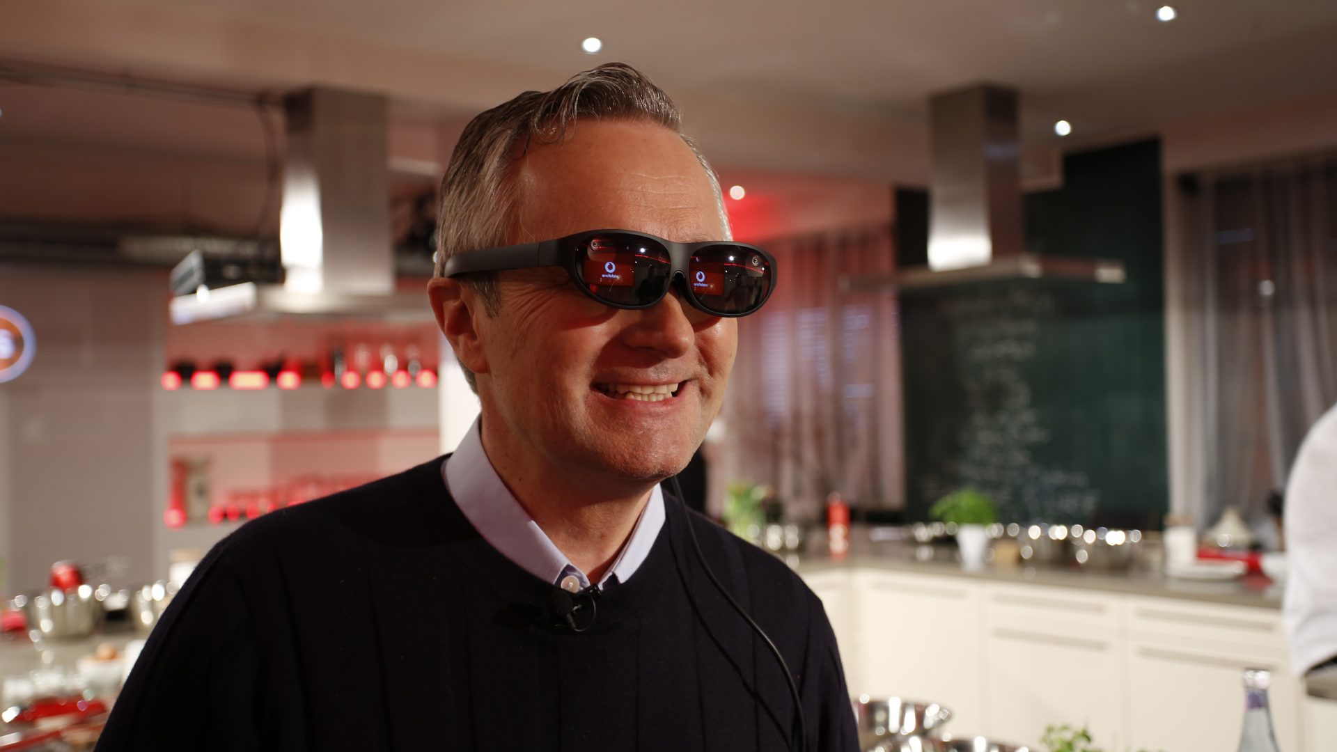 Vodafone CEO Hannes Ametsreiter steht in einer modernen Küche, die mit stilvollen Elementen wie Edelstahlgeräten und Kräutertöpfen ausgestattet ist. Im Hintergrund sind Küchenschränke, eine große Arbeitsfläche und eine Tafel zu sehen. Die Beleuchtung ist warm und schafft eine einladende Atmosphäre.