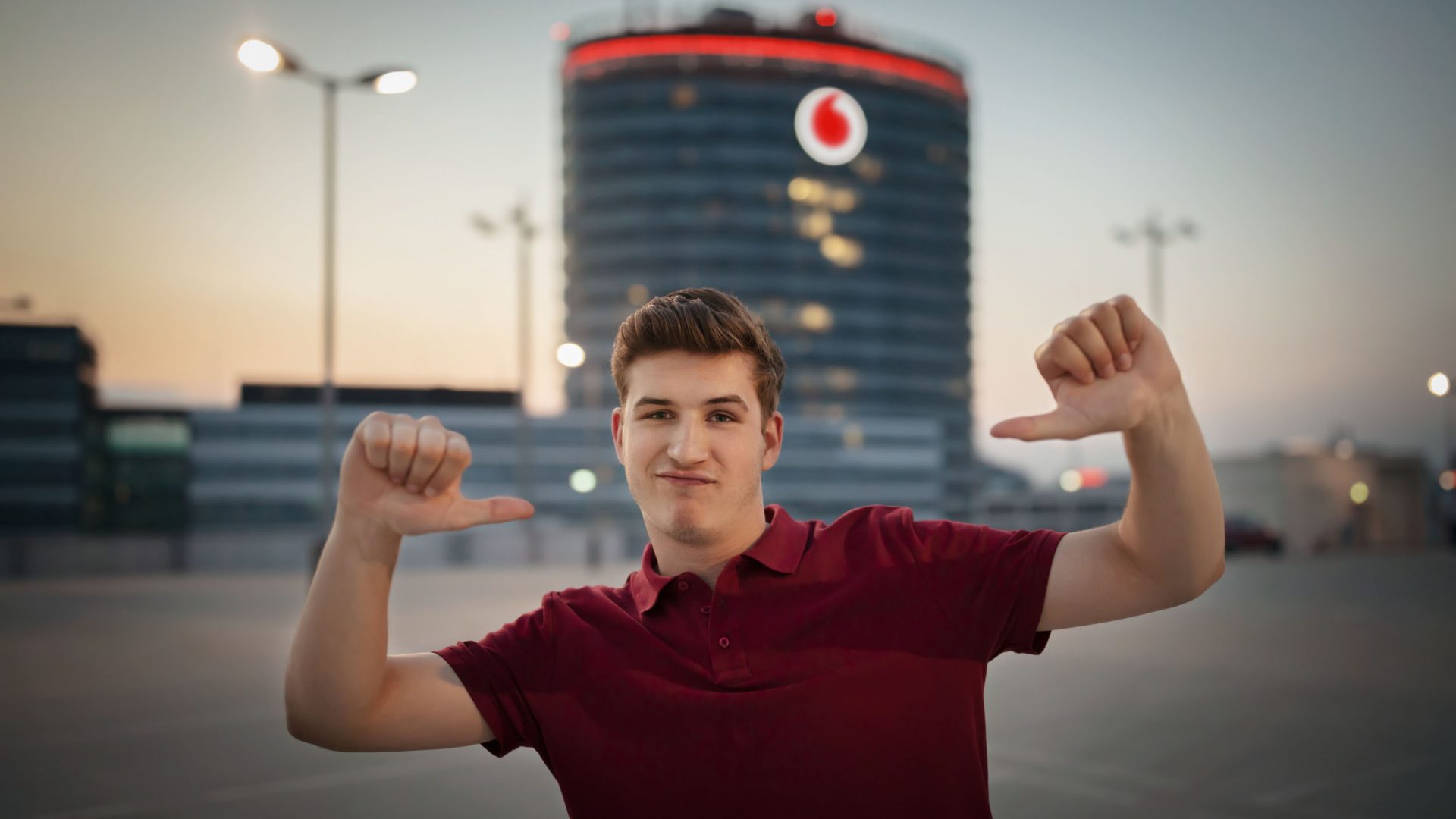 Eine Person steht auf einem Parkplatz und zeigt mit beiden Daumen auf sich selbst. Im Hintergrund ist der Vodafone Campus in Düsseldorf zu sehen, beleuchtet in der Dämmerung. 