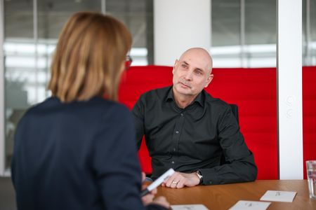 Zwei Personen sitzen in einem modernen, roten Sitzbereich. Eine Person hält einen Stift in der Hand, während sie sich mit der anderen Person unterhält. Der Raum ist hell und stilvoll eingerichtet.