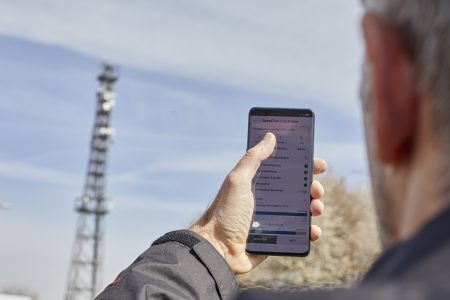 Ein Mann hält ein Smartphone in der Hand und betrachtet den Bildschirm, während er vor einem Mobilfunkmast steht. Der Fokus liegt auf dem Telefon, das Informationen zur Netzabdeckung anzeigt. Der Mobilfunkmast ist im Hintergrund sichtbar, und der Himmel ist klar mit einigen Wolken.