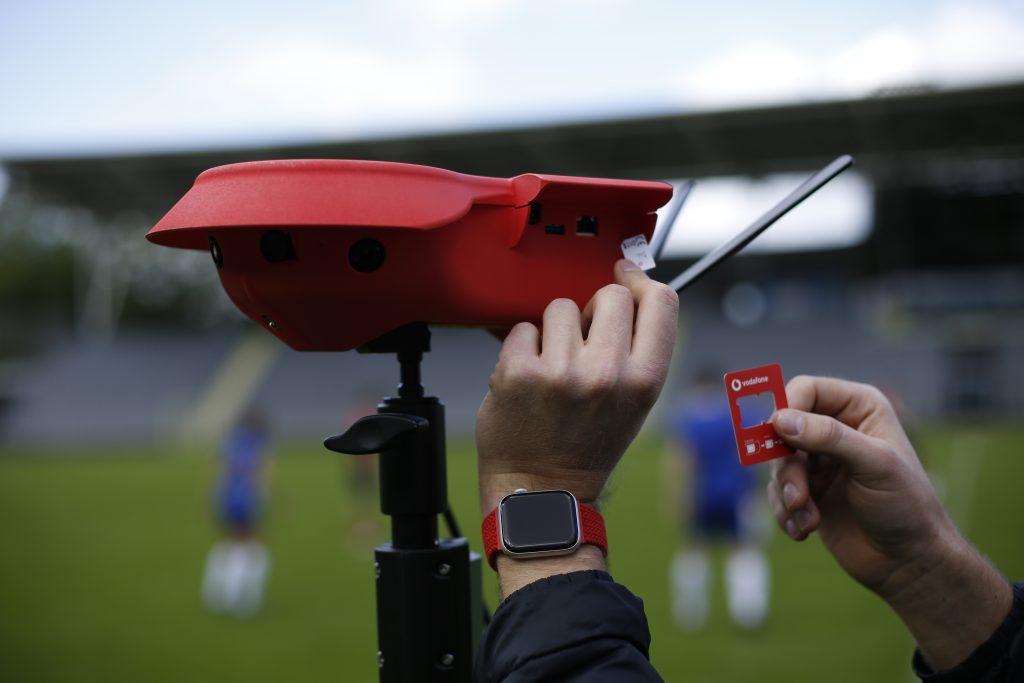 Eine Person installiert eine rote SIM-Karte in ein rotes Gerät mit Antennen, das auf einem Stativ montiert ist. Im Hintergrund sind unscharf Fußballspieler und leere Tribünen zu sehen.

