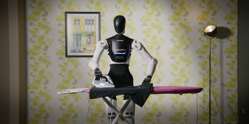 Ein humanoider Roboter mit schwarzem Torso und weißen Armen steht vor einem Bügelbrett und bügelt ein schwarzes Kleidungsstück. Der Roboter hält ein Bügeleisen in der rechten Hand, während die linke das Stoffstück festhält. Im Hintergrund befindet sich eine Wand mit gelb-grünem Blumenmuster, ein gerahmtes Bild an der Wand zeigt bunte Gebäude und ein Fenster. Rechts steht ein Studio- oder Scheinwerferlicht auf einem Stativ.