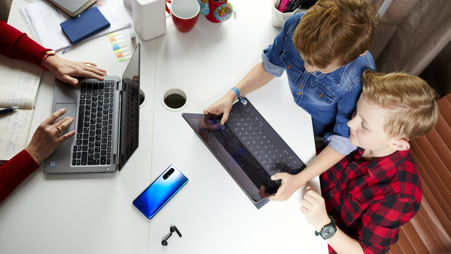Drei Personen an einem weißen Tisch. Links arbeitet eine Person an einem Laptop, während rechts zwei Kinder gemeinsam auf ein Tablet schauen. Auf dem Tisch liegen ein Laptop, Notizbücher, ein Smartphone, Ohrhörer, Stifte in einem Becher und ein roter Becher.