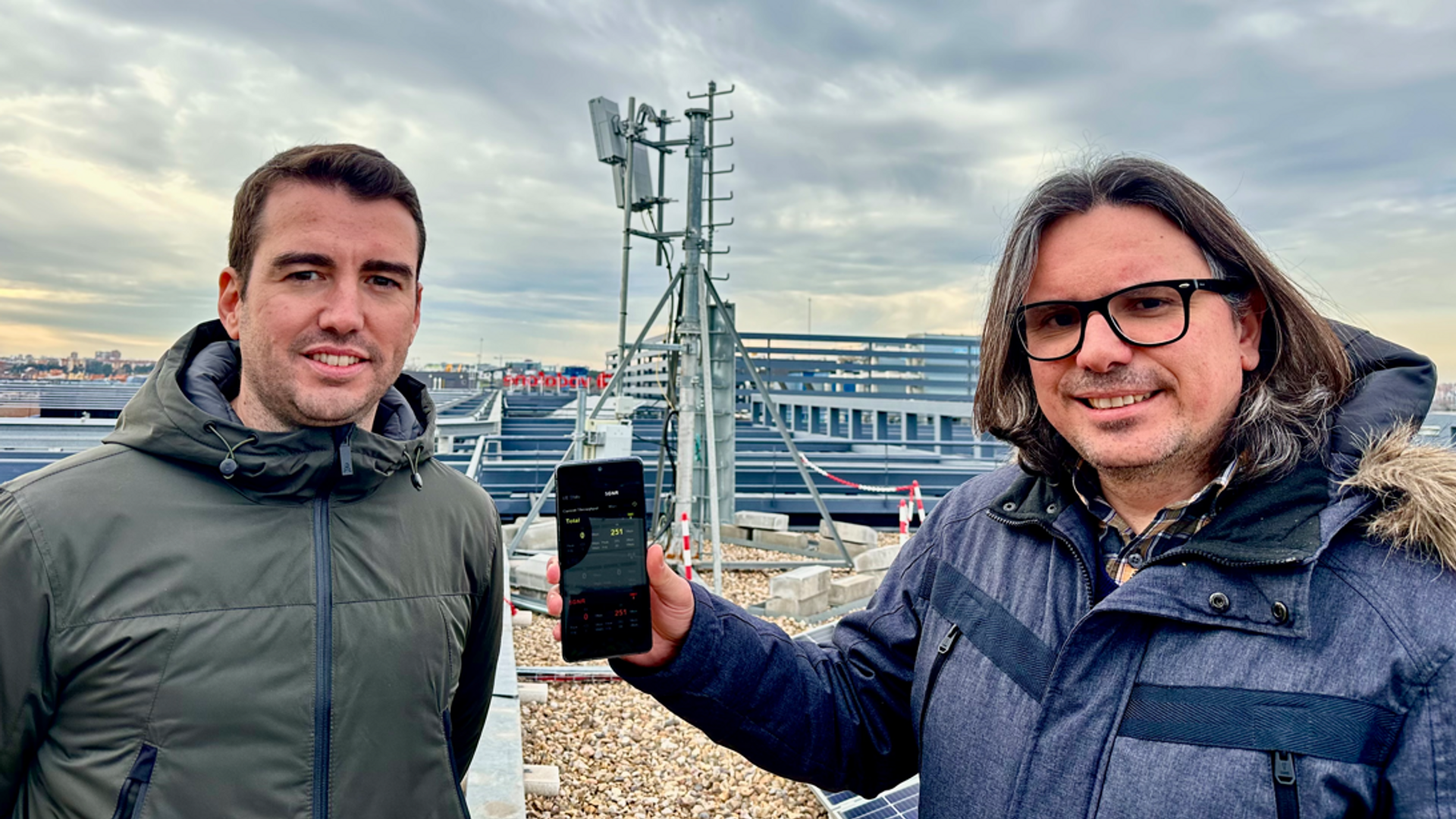 Zwei Personen stehen auf einem Dach mit einer Mobilfunkantenne im Hintergrund. Eine Person hält ein Smartphone in der Hand, das möglicherweise zur Messung oder Demonstration von Signalstärke verwendet wird. Der Himmel ist bewölkt, und im Hintergrund sind städtische Gebäude zu sehen, die eine urbane Umgebung schaffen.