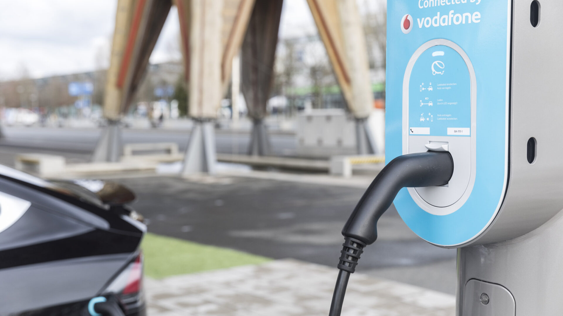 Ladestation mit eingestecktem E-Auto-Ladekabel, im Hintergrund ein geparktes Elektroauto.