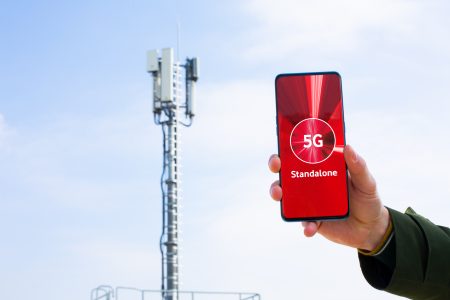 Eine Hand hält ein Smartphone, dessen Bildschirm die Worte "5G Standalone" auf einem roten Hintergrund zeigt. Im Hintergrund ist ein Mobilfunkmast gegen einen klaren blauen Himmel zu sehen.