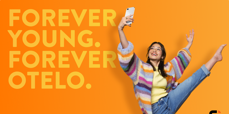 Werbegrafik mit orangefarbenem Hintergrund. Links steht in großen, gelben Buchstaben: „FOREVER YOUNG. FOREVER OTELO.“ Rechts daneben posiert eine junge Person in bunter Strickjacke, gelbem Shirt und Jeans, macht ein Selfie mit dem Smartphone und streckt ein Bein dynamisch in die Luft.