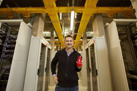 Guido Weissbrich, Chef-Netzplaner bei Vodafone Deutschland, steht in einem modernen Rechenzentrum zwischen mehreren Server-Racks. Er trägt eine schwarze Jacke und hält ein rotes Vodafone-Produkt in der Hand. Über ihm verlaufen gelbe Kabelkanäle an der Decke, die die technische Infrastruktur des Raumes betonen. Die Umgebung ist hell und gut organisiert.