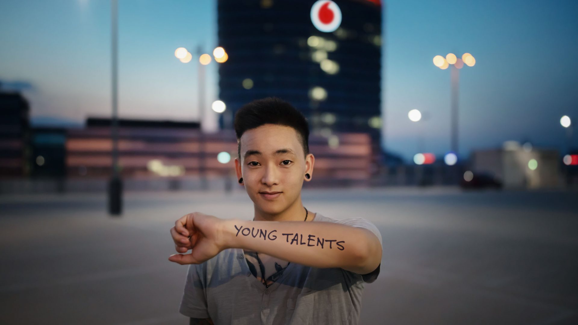 Eine Person steht auf einem Parkplatz und hält ihren Arm ausgestreckt, auf dem "YOUNG TALENTS" geschrieben steht. Im Hintergrund ist der Vodafone Campus in Düsseldorf zu sehen, beleuchtet in der Dämmerung.