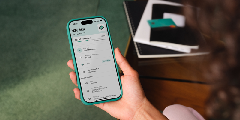 Eine Hand hält ein Smartphone, auf dessen Bildschirm eine App mit dem Titel "N26 SIM" geöffnet ist. Die App zeigt Informationen zu einem Mobilfunkvertrag, einschließlich des verfügbaren Datenvolumens und der aktuellen Nutzung. Im Hintergrund liegen Notizbücher und ein Stift auf einem Tisch, was eine alltägliche, produktive Umgebung suggeriert.